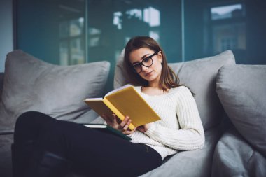 Genç, ciddi bir kadın, gözlüklü, gözlüklü, puf mindere uzanmış, dinlenirken ilginç bir kitap okuyor.
