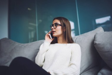 Yumuşak koltukta otururken gözlerini kaçıran ve arama yapmak için cep telefonunu kullanan düşünceli bir kadının alçak açısı.