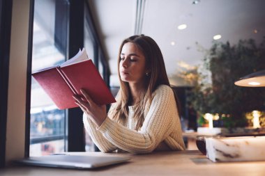 Gündüz vakti kafeteryada vakit geçirirken elini kaldırmış ve not defterinden notlar okuyan düşünceli bir kız öğrenci.
