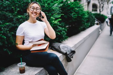 Eğitim kitaplarındaki neşeli bayan öğrenci uluslararası cep telefonu konuşmaları sırasında organizasyon planlayıcısını tartışıyor, gözlüklü mutlu Çinli kadın arkadaşça sohbetten zevk alıyor.