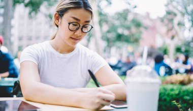 Optik gözlük takan Asyalı bayan öğrenci açık havada öğrenim görürken eğitim kitabına notlar yazıyor. Zeki Japon kadın sınavlara hazırlanırken bilgi okuyor.