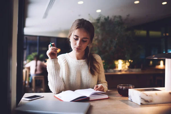 暖かいセーターを着た茶色の髪をした女性の学生がスマートフォンと飲み物のカップでテーブルに座って 昼間のカフェでノートを読む — ストック写真