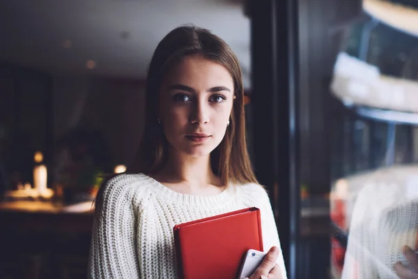 ノートパッドと携帯電話で立っている間にカメラを見て長い髪を持つカジュアルな服の若い気配りの女性学生 — ストック写真