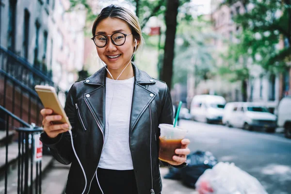 Elektronik kulaklıklı mutlu Çinli kullanıcının elinde akıllı telefon cihazı, elinde kahve içeceği ve kameraya gülümseyen cep telefonlu güzel hippi kız müzik dinlemekten hoşlanıyor.