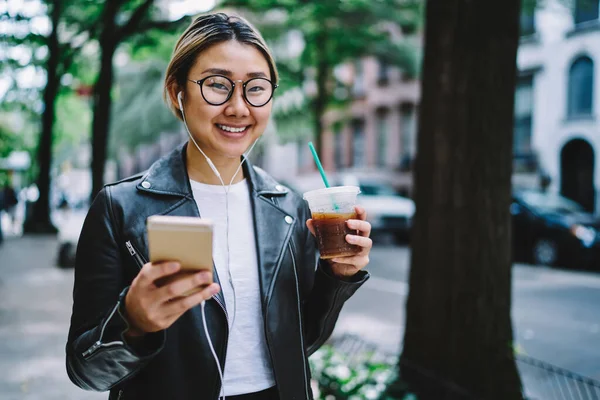 Elektronik kulaklıklı mutlu Çinli kullanıcının elinde akıllı telefon cihazı ve elinde kahve içeceği kameraya gülümseyen cep telefonlu neşeli hippi kız müzik dinlemekten zevk alıyor.