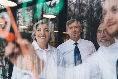 Modern ofiste yeni bir proje üzerinde çalışırken şeffaf tahtaya notlar yazan neşeli iş ortaklarıyla dolu bir cam aracılığıyla.