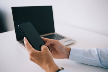 Yukarıdan gelen bilgilere göre, isimsiz bir erkek girişimci, iş sırasında ahşap masada dizüstü bilgisayarla oturuyor ve cep telefonuyla mesajlaşıyor.