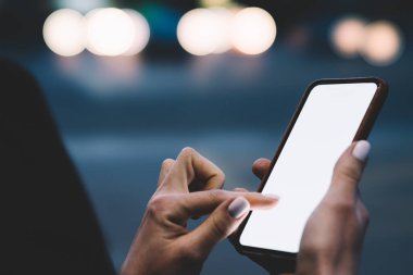 Akşamları aydınlık şehir sokağında vakit geçirirken siyah kazaklı, akıllı telefonlu, ekili, isimsiz bir kadının yan görüntüsü