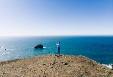 Mavi okyanusun kenarındaki tepede yaz günü açık gökyüzüne karşı duran uzun saçlı, tanınmayan bir kadın vücudu.