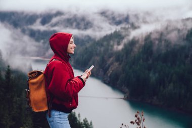 Sırt çantalı, akıllı telefonlu, sisli orman ve nehir manzarasını keşfederken sıcacık kıyafetli gülümseyen kadın gezginin yan görüntüsü.