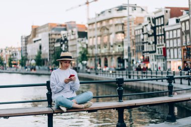 Amsterdam Hollanda 'da tatil sırasında Lotus bankında poz verirken gündelik kıyafetleriyle modern cep telefonuna göz atan odaklanmış kadın blogcu tam boy