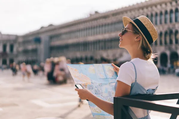 紙の地図で観光しながら ダウンタウンの広場の真ん中に立つ麦藁帽子の楽観的な女性の側面図 — ストック写真