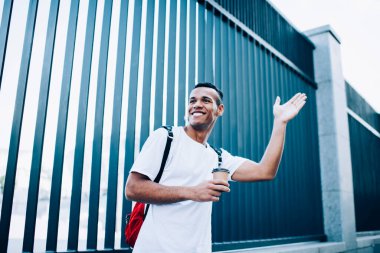 Elinde kağıt bardakla metal kapıların yanında duran ve sokaktaki arkadaşa el sallayan neşeli Afro-Amerikan erkek.