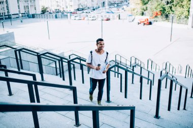 Yüksek açılı, kısa saçlı, netbook ve kahve taşıyan, yaz günü metal tırabzanlarla merdiven boyunca yürüyen mutlu, Afro-Amerikan bir erkek.