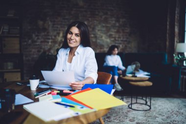 Beyaz gömlekli neşeli, genç esmer, yuvarlak bir masada oturuyor. Yanında kâğıtlar ve kırtasiye kağıtları var. Bulanık iş arkadaşının yanında.