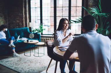 Gündelik beyaz gömlekler giyen gülümseyen genç iş arkadaşları modern yaratıcı iş yerinde birlikte çalışırken masada toplanıp iş stratejisi hakkında konuşuyorlar.