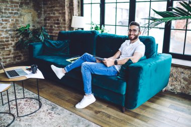 Yan görünüş, kot pantolon ve beyaz gömlekli gülümseyen genç adam rahat kanepeye uzanıyor ve çağdaş iç mekanlarda tablet kullanıyor.