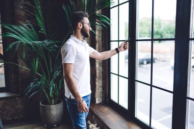 Gündüz vakti ayakta duran ve büyük pencereden dışarı bakan sıradan giyinmiş, şık gözlükler takan sakallı genç adamın yan görüntüsü.