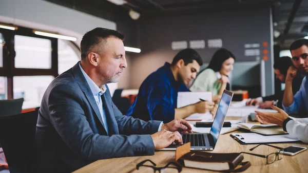 Olgun erkek şirket müdürü modern dizüstü bilgisayarı kullanarak finansal web sitesinde bilgi alışverişi yapıyor, iş adamı bankacılık yaparken iş arkadaşları iş birliği yapıyor.