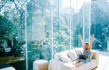 Heyecanlı, uzaktan kumandalı, netbook kullanan, kollarını kaldıran, modern çalışma sahasında günışığında camdan duvarları olan bir kadın.