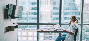 Sıradan kıyafetlerde kadın uzmanın bakışları başka tarafa bakıyor ve çeneye dokunuyor dizüstü bilgisayar ve belgelerle masada oturuyor.