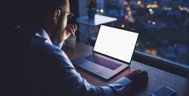Resmi kıyafetli ve gözlüklü isimsiz bir erkek işçi gece boyunca modern ofiste iş projesi üzerinde çalışırken dikkatle bilgisayar ekranına bakıyor.