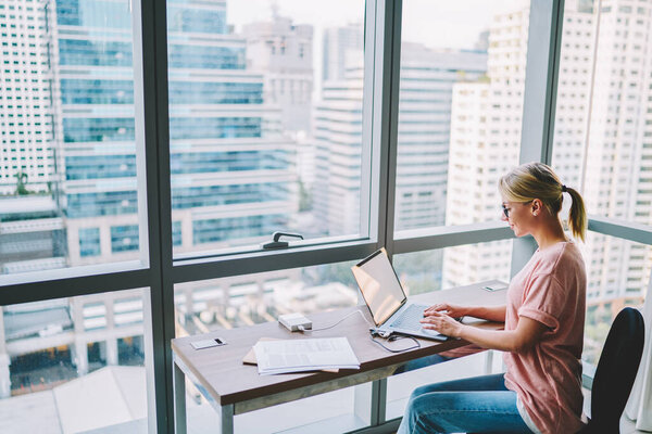 Боковой вид улыбающейся женщины в повседневной одежде и очках, сидящей на ноутбуке во время работы над проектом в просторном современном рабочем пространстве с видом на город в дневное время