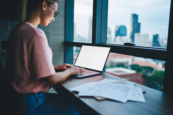 Вид сбоку на даму в повседневном наряде и солнцезащитных очках, использующую ноутбук для работы и анализа документов, глядя на экран в современном офисе рядом с большим окном