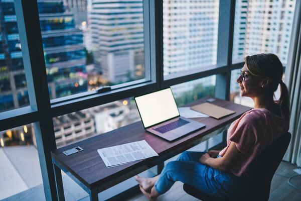 Высокий угол улыбки сотрудницы смотрит в сторону сидя за столом с ноутбуком и документами во время работы в офисе в вечернее время