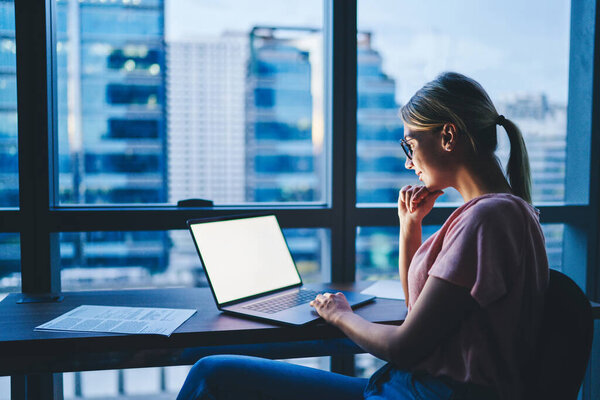 Вид сбоку молодой концентрированной женщины, касающейся подбородка, сидя за столом, просматривая ноутбук во время работы вечером в офисном кабинете