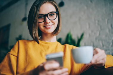 Kafenin içinde boş zamanlarında kameraya gülümseyen modern cep telefonu cihazıyla ve elinde çay fincanıyla mutlu Kafkasyalı kullanıcının portresi, cep telefonu tutan güzel hippi blogcu.