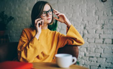 Göz düzeltmesi için optik gözlüklü düşünceli hippi kız kahve dükkanında boş zamanlarında danışmanlık akıllı telefon sohbeti yapıyor düşünceli bayan müşteri cep telefonu bağlantılarını tartışıyor.