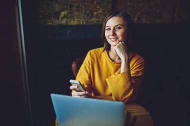 Modern cep telefonu cihazıyla tasasız kadın blogcu portresi. Kapalı mekanda cep telefonu ve dizüstü bilgisayarla iş birliği yaparken kameraya bakıyor.