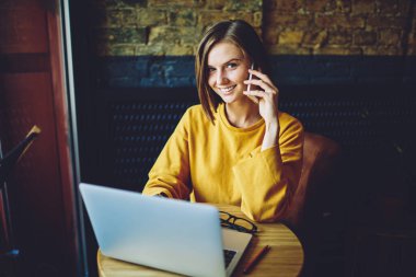 Kafedeki cep telefonu konuşmaları sırasında kameraya gülümseyen neşeli bir kadının portresi, mutlu hippi kız netbook programlama hakkında konuşmak için arkadaşça cep telefonuna sahip.