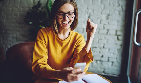 Cheerful female in classic eyeglasses happy with received email message using cafe internet connection on cellular technology, smiling blogger reading online reviews browsed on smartphone device