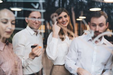 Pencere pozitifliği ile resmi meslektaşlar modern ofiste birlikte ayakta durarak ve gülerek başlangıç planını cama çizerken.