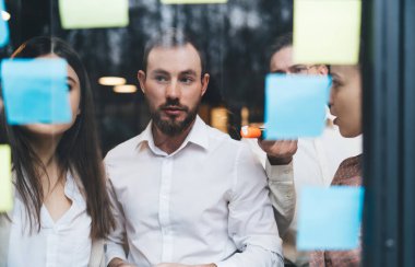 Bir grup ciddi iş ortağı bir arada duruyor ve iş sırasında konferans odasında toplantı yaparken yapışkan notlara bakıyor.