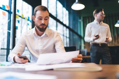 Odaklanmış genç erkek çalışanlar resmi kıyafet içinde masa başında oturmuş evrakları okuyor ve ofiste çalışırken cep telefonlarını karıştırıyorlar.