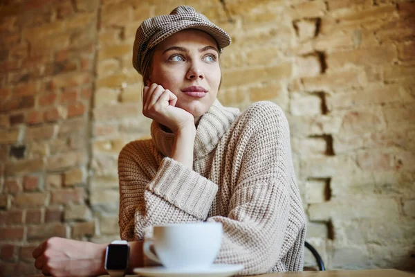 おしゃれなセーター姿のシリアスな女性の低角度離れてコーヒーを飲みながらカフェでテーブルに座って手にもたれて見て — ストック写真