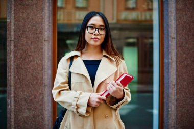 Uzun saçlı ve gözlüklü genç, düşünceli Asyalı kız öğrenci şehir sokaklarında planlamacıyla kameraya bakıyor.