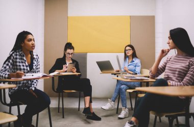 Modern sınıfta ahşap masalarda oturup öğretmenlerini beklerken birbirlerine bakan çok kültürlü genç bayan öğrenciler.