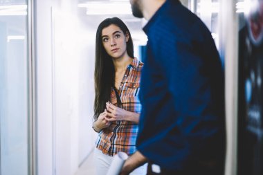 Uzun saçlı, günlük giysiler içinde genç bir bayan yönetici ofis koridorundaki bulanık arka plandaki yeni proje hakkında meslektaşıyla konuşuyor.