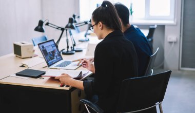 Not defteri ve masa lambasıyla masada oturan ve modern ofiste dizüstü bilgisayar üzerinde çalışan meslektaşların arka planı
