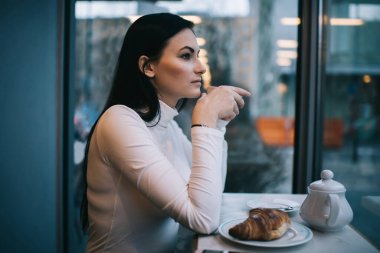 Uzun siyah saçlı, gündelik kıyafetleri olan genç bir kadının ayçöreğiyle masaya oturması ve kahve içmesi.