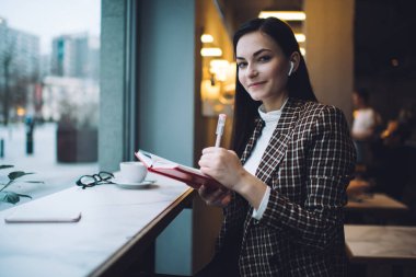 Rahat bir kafede organizatörden notlar alırken şık giyinen, sıcak içecekle gülümseyen neşeli genç bir kadın.