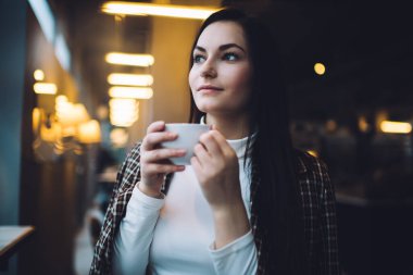 Uzun siyah saçlı, ekose ceket giyen, modern kafede vakit geçiren ve çay içen pozitif genç bir kadın.