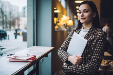 Zarif kıyafetli genç bir kadın elinde bilgisayarla elinde not defteri ve telefon ile gün ışığında kafedeki panonun yanında kameraya bakıyor.