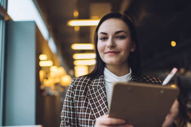 Kareli ceketli, uzun siyah saçlı, kameraya bakan ve kafedeki panoya not alan genç bir kadın.