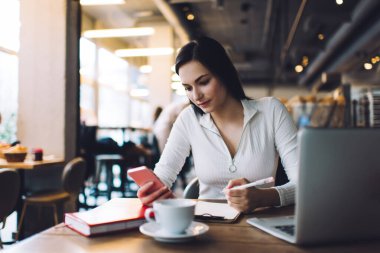 Modern kafeteryadaki masada oturmuş, yeni bir proje üzerinde çalışırken akıllı telefonları karıştıran, gündelik kıyafetli genç bayan uzaktan çalışan.