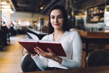 Moda kıyafetleri içinde hoş, genç bir bayan rahat modern kafede otururken ilginç bir kitap okuyor.